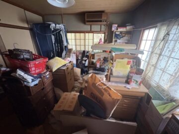 IMG_0067 | あつまる君 A cluttered room filled with stacked boxes, clothes, and miscellaneous items scattered across furniture and floors, with sunlight from the windows.