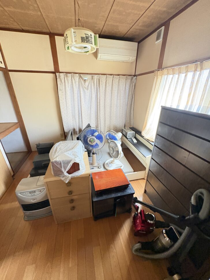 Small, cluttered room with cardboard boxes and a dresser atop a wooden floor; two blue oscillating fans sit on a table in front of a curtained window, with a vacuum cleaner and other appliances scattered nearby.