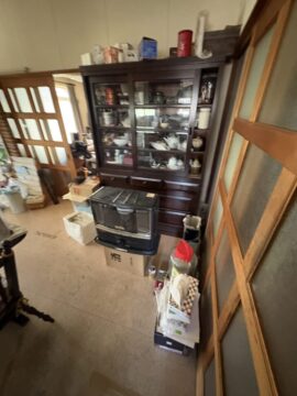 A cluttered living room corner with a dark wooden china cabinet filled with dishes and glassware, a freestanding heater in front, and stacked boxes and clutter nearby.