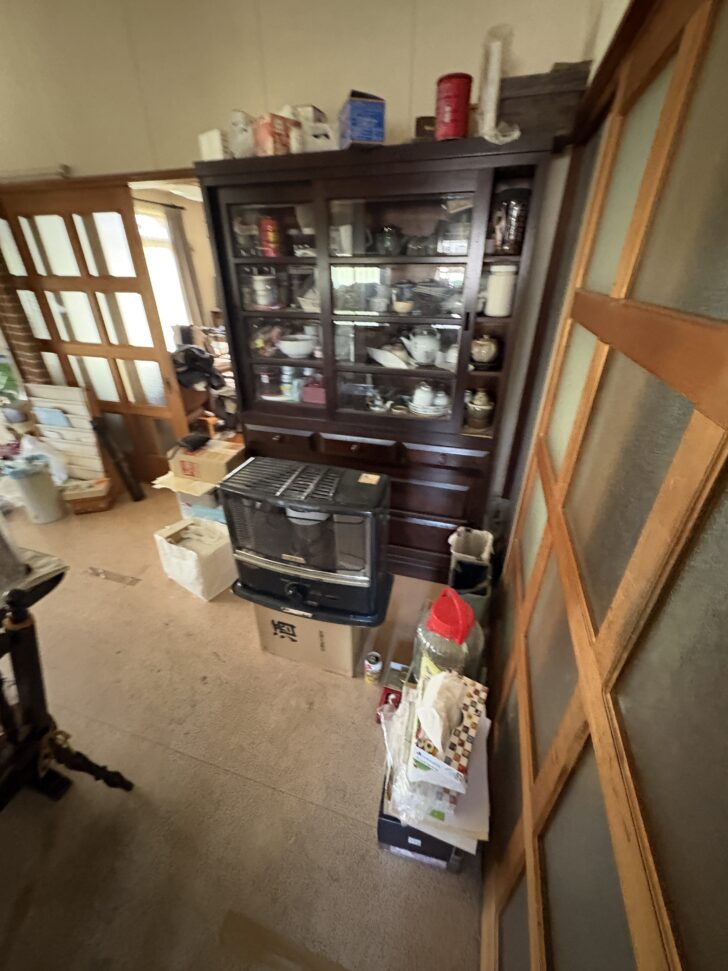 A cluttered living room corner with a dark wooden china cabinet filled with dishes and glassware, a freestanding heater in front, and stacked boxes and clutter nearby.