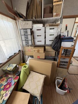 Cluttered storage room with a metal shelving unit and stacked boxes, baskets, and miscellaneous items on shelves and floor window side light.