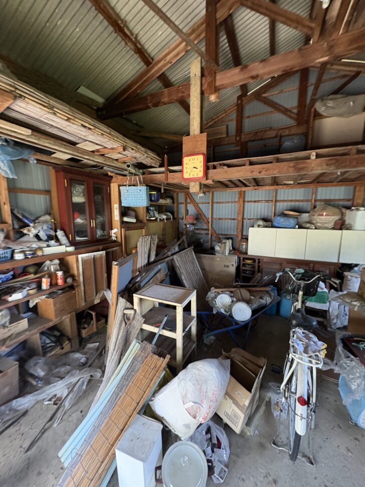 Cluttered wooden shed workspace with stacked lumber, shelves, and a bicycle among scattered tools and boxes, a yellow clock overhead.