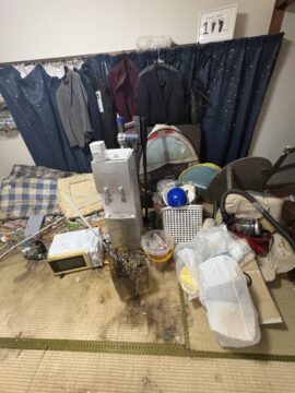 Cluttered room with clothes hanging on a rack, a metal machine, tangled cords, and assorted bags and tools strewn on a tatami mat floor.