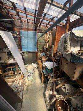 Cluttered shed interior with shelves, tools, and scraps scattered on the floor under a translucent corrugated roof with sunlight.