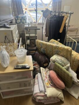 A cluttered room with a clothing rack, sofa, cushions and blankets piled on the floor, and a window in the background.