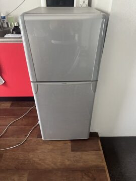 Tall silver refrigerator/freezer unit in a kitchen, with a red cabinet to the left and cords on the wooden floor.