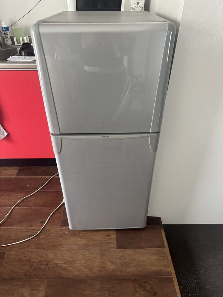 Tall silver refrigerator/freezer unit in a kitchen, with a red cabinet to the left and cords on the wooden floor.
